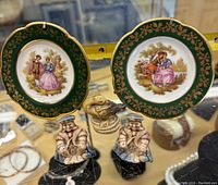 Two Limoge miniature decorative plates with green and gold floral border, depicting 18th-century style couples in the center. Three Wade Whimsy ceramic figurines in front, showing small characters seated on black bases.