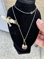 Enamel bird pin with outstretched wings and sterling silver chain with bird nest pendant displayed on black stand.