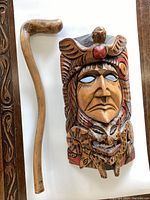 Carved wooden mask with shillelagh walking stick on the left, showing natural wood finish and painted details on mask.