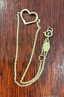 Gold heart-shaped charm attached to fine gold chain with spring ring clasp, laid flat on wooden surface.