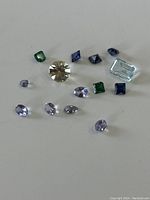 Loose mixed gemstones scattered on white background, showing variety of colors and cuts including a prominent round clear stone, rectangular light blue stone, and green and blue square and oval stones.