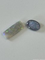 Two opal cabochon gemstones laid flat showing their polished tops, one rectangular with green and purple iridescence, the other oval with dark tones and color play.