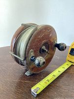Front angle side view of wooden fishing reel with white fishing line and two worn black and wooden handles adjacent to measuring tape showing size.