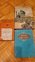 Three books laid on parquet floor: The Arabian Nights with worn illustrated dust jacket, Arabian Love Tales in teal hardcover, and a third book with orange cover.