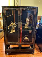 Black lacquer cabinet front with enameled figures and gold painted decorative borders, showing two doors and red latches.