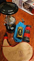 Set showing black Coleman lantern, leather wine flask, yellow candle lantern in blue pouch, red candle lantern on wood floor, and boxed Coleman lantern in background
