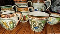 Close-up of several Adams ceramic jugs and cups depicting colorful scenes and quotes from Dickens and Shakespeare with detailed orange patterned rims on a wooden surface.