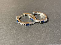 Pair of silver hoop earrings with rectangular blue stones and clear accent stones laid flat on black background.