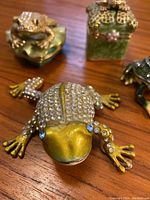 Close up of a frog-shaped jewelry box decorated with clear rhinestones, gold and green paint, and blue rhinestone eyes