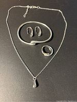 Top-down view of the silver-tone jewelry set including necklace, ring, bracelet, and earrings on a black surface