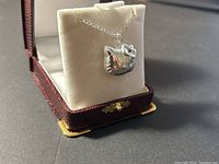 Kitty cat silver-tone locket pendant necklace displayed on cream velvet holder inside a brown textured jewelry box with gold corner accents.