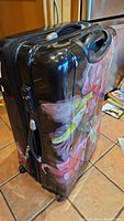 Side angle view of the large black hard-shell luggage featuring colorful hummingbirds and pink flowers. Shows top handle, side scuffs, and wheels.