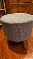 Side-angle view showing round gray jute fabric upholstered stool resting on wood floor with wooden dresser in background.