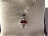 Close-up photo of the Pokemon Pokeball pendant on a silver-tone chain displayed on cream-colored cushion, showing the red and clear enamel details and central gemstone.