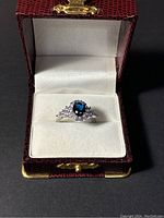 Front view of sterling silver ring with blue center stone and clear stones inside red jewelry box with white interior.