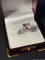 Ring in a red textured box with white cushioned interior, showing the heart-shaped pink stone and clear accent stones.