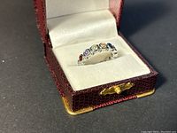 Ring in a red textured case with white cushioned interior showing multi-colored gemstones on silver band from one angle.