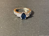 Front view of sterling silver ring showing blue oval center stone and clear side stones with engraved band