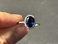 Close-up of the ring held between fingers showing oval dark blue gemstone surrounded by small clear stones on sterling silver band