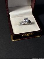 Ring set displayed inside a burgundy textured ring box with white cushioned interior, showing the solitaire ring with large center cubic zirconia stone and pavé stones on band