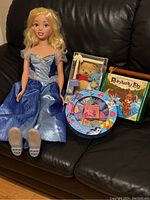 Cinderella doll seated on black chair, dressed in blue and silver gown, with blonde hair, next to Disney My First Pooh bear box, Runaway Log game, and My Little Pony game