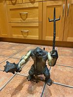 Front view of Cave Troll action figure standing on tile floor holding two weapons, showing full figure and detail of arms, face, and torso