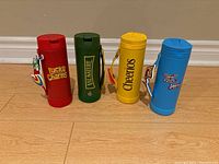 Four cereal themed water bottles in red, green, yellow, and blue, standing upright showing front logos and attached charms.