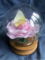 Full view of the glass globe containing the yellow butterfly perched on the pink rose with green leaves, mounted on a wooden base.