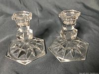 Pair of clear glass candle holders featuring 3-inch wide hexagonal bases with starburst patterns, photographed against a dark background.