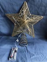 Photo of gold-colored wire star shaped Christmas tree topper with spiral base and attached battery pack with AA batteries.