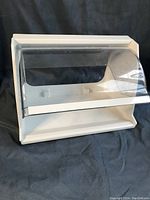 White plastic countertop bread bin with clear rotating front cover on dark fabric background.