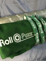 Green felt Roll O Puzzle mat rolled up with three green printed ties and gray cylindrical storage tube behind.