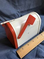 Metal mailbox coin bank shown next to a ruler for size reference, with red plastic flag up and metal latch on top.