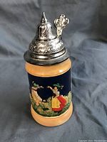 Front view of mini beer stein showing colorful traditional figure artwork on dark blue background with beige top and base, and ornate pewter lid.