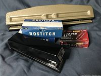 Metal three-hole punch, black stapler, Bostitch staples and Acco paper clips boxes displayed on fabric surface.