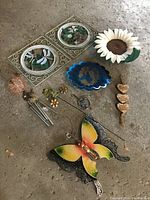 Photo shows 7 items: metal dragonfly wall art, sunflower wall decor, butterfly spinner, wind chime with colorful dangles, blue leaf ceramic dish, decorative floral stake, and hanging hearts decoration.