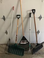 Five outdoor maintenance tools including snow shovels and rakes leaning against a garage door.