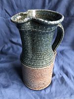Front and side view of handcrafted pottery pitcher showing its textured green and brown glaze and handle.