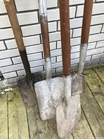 Four assorted garden shovels with wooden handles leaning against a brick wall, showing signs of wear and rust on the metal blades.
