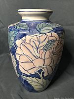 Photo of a tall ceramic vase with blue background and pink flowers with green leaves, showing the full front view and neck rim.