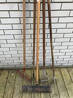 Photo shows five gardening tools leaning against white brick wall on wooden ground including two rakes, pitchfork, garden fork, and broom, all with wooden handles and metal functional ends.