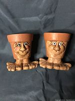 Top view of two whimsical terracotta plant holders with faces and feet, shown against a dark fabric background.