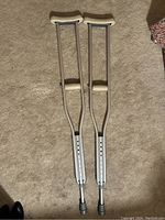 Pair of silver aluminum medical crutches lying flat on carpet, showing full length with padded underarm grips and hand grips.