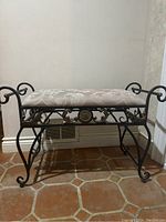 Front view of the decorative metal framed bench with upholstered seat showing black metal with gold floral medallion and scrollwork.