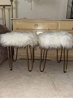 Pair of two white faux fur stools with gold hairpin legs, positioned side by side in front of wooden furniture.