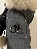 Close-up of left upper sleeve pocket with black glossy Moose Knuckle logo and silver rivets on dark fabric.