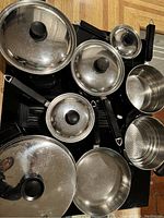 Top-down photo showing the collection of stainless steel pots, pans, and lids with black handles and knobs arranged on a kitchen surface.