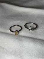 Photo of two delicate sterling silver rings lying on a fabric surface; one with a yellow citrine stone, the other with a clear stone and bead accents.