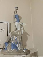 Side view of the vintage porcelain figurine showing the woman in blue top and white skirt standing beside a greyhound dog on a textured base.