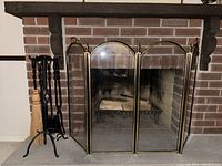 Fireplace screen and tool set in front of fireplace showing the screen's brass trim and three panels and the iron tool stand holding tools.